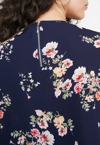 Robe florale bleu marine en tissu doux, ornée de fleurs roses et blanches. Comprend une fermeture éclair dans le dos et une surface lisse et texturée.