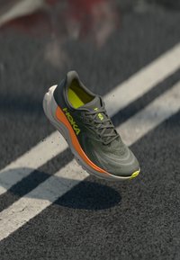 Laufschuh in Grün mit orangefarbenen und gelben Akzenten, in der Luft �über einer Asphaltstraße mit weißen Fahrbahnmarkierungen.