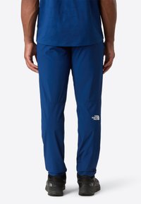 Osoba v modrých kalhotách North Face a černých turistických botách, stojící se svýma rukama u těla proti jednolitému pozadí.