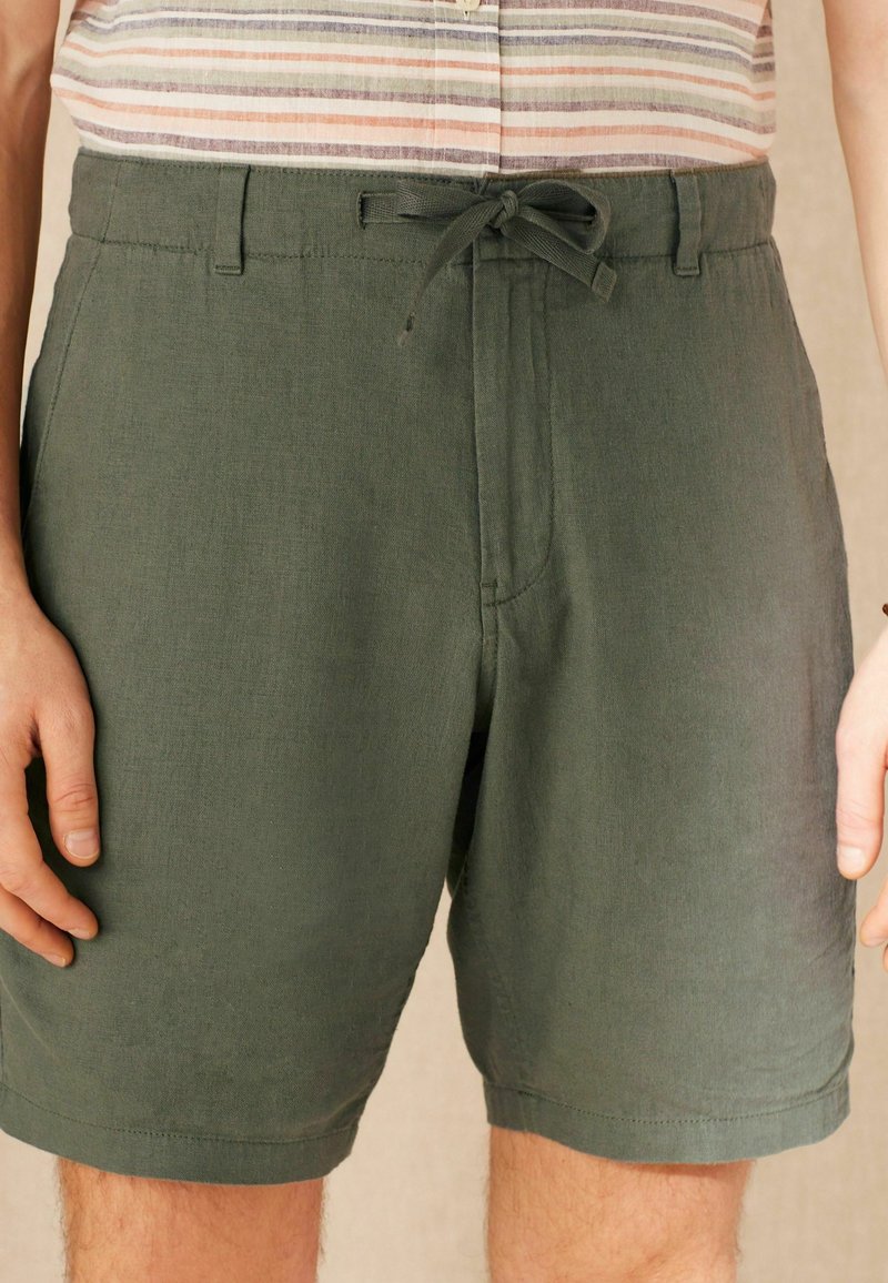 Pantaloni corti in lino verdi con vita regolabile, tasche frontali e vestibilità comoda, caratterizzati da una texture liscia e sottili dettagli di cucitura.