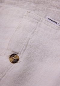 Witte stof met een gestructureerde weving, voorzien van een tan kleurige knop en een klein label met de tekst "Lindbergh" in de hoek.