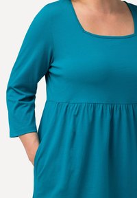 Tyrkysové šaty se čtvercovým výstřihem, tříčtvrtečními rukávy a sbíraným pasem s kapsami. Měkká, lehká tkanina s hladkým povrchem.
