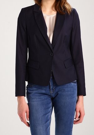 Femme portant un blazer noir ajusté avec un seul bouton, un chemisier clair et un jean bleu, debout avec les bras détendus.