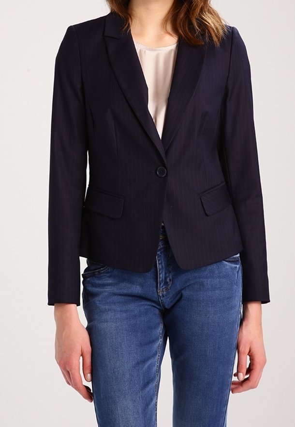 Femme portant un blazer noir ajusté avec un seul bouton, un chemisier clair et un jean bleu, debout avec les bras détendus.