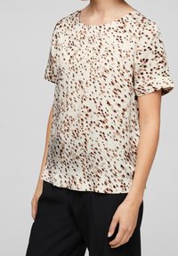 Blusa a maniche corte color beige chiaro con motivo maculato marrone, dotata di scollatura rotonda e vestibilità morbida. Tessuto liscio, con caduta delicata.