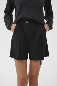 Persona con pantalones cortos negros con cinturón y un suéter de manga larga gris oscuro, con las manos en los bolsillos, mostrado desde mitad del muslo hasta la cintura.