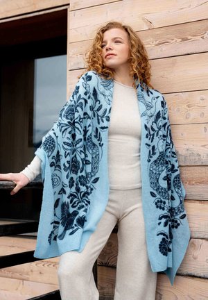 Sciarpa azzurro chiaro con motivi floreali e uccelli blu scuri, realizzata in materiale morbido, drappeggiata sulle spalle, che contrasta con un outfit beige.