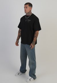 Camiseta de algodón negra con mangas cortas y un logo gráfico en el pecho, combinada con jeans de mezclilla azul claro y zapatillas blancas.