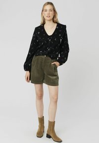 Blouse noire à motifs floraux avec col à volants, associée à un short en velours côtelé olive et des bottines beiges. Les accessoires comprennent des colliers superposés et des boucles d'oreilles.