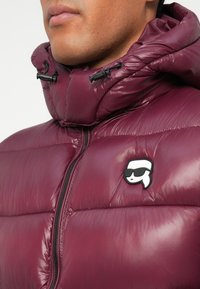 Burgundska puffer jakna s sjajnom teksturom, koja ima uspravni ovratnik, zatvarač sprijeda i kontrastni crni logo na prsima.