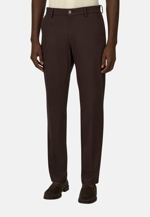 Homme portant un pantalon ajusté marron foncé et des chaussures marron foncé assorties, debout avec les mains détendues le long du corps.