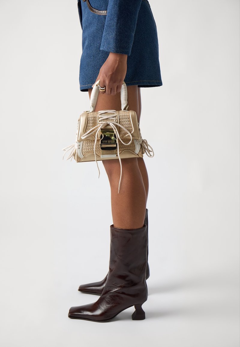 Personne en jupe en denim bleu tenant un sac à main tissé beige et blanc, portant des bottes en cuir marron foncé à talons mi-mollet sur fond blanc.