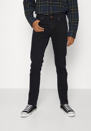 Homme portant un jean slim noir, des baskets noir et blanc, et une chemise à carreaux vert foncé à manches longues, debout avec une main dans la poche.