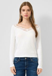 Junge Frau trägt ein weißes Langarmshirt mit Spitzenbesatz am Ausschnitt und blaue Jeans, steht vor einem schlichten hellen Hintergrund.