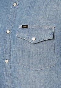 Vaaleansininen farkkupaita, jossa on terävä tasku, hopeiset napit ja pieni musta etiketti valkoisella tekstillä. Sileä pinta ja rento malli.