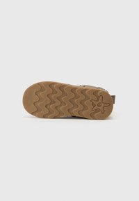 Semelle en caoutchouc avec une texture en zigzag, couleur marron, et un logo en étoile. Tige beige avec un design durable et une sangle ajustable pour un meilleur maintien.