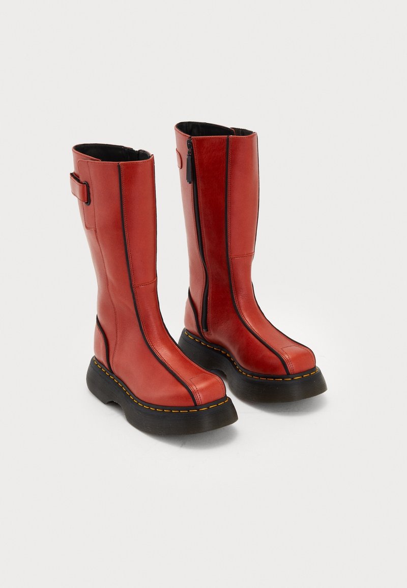 Bottes hautes rouges en cuir avec semelles épaisses noires, coutures jaunes, fermetures éclair latérales et sangles réglables en haut, présentées sur un fond blanc.