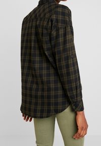 Chemise à carreaux noire et olive, avec un col, des poignets boutonnés et un ourlet arrondi, fabriquée en tissu doux, avec une coupe décontractée.