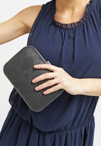 Musta tekstuuriga klamberkott, millel on sisalikusarnane muster, ristkülikukujuline vorm ja metallist sulgur, hoitakse käes tumesinise pluusi taustal.