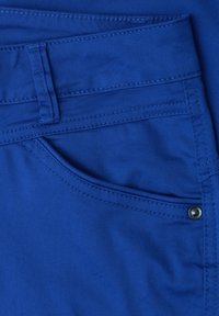 Gros plan d'une poche de pantalon en tissu bleu avec des coutures visibles, un passant de ceinture et un détail de rivet métallique sur la couture latérale.