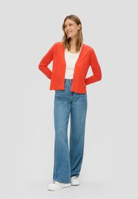 Cardigan lavorato a maglia rosso con maniche lunghe, aperto davanti, indossato su un top bianco e jeans a gamba larga azzurri chiari, abbinato a sneakers bianche.
