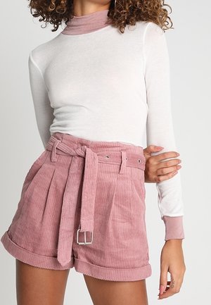 Personne portant un haut blanc à manches longues et un short en velours côtelé rose taille haute avec une ceinture en tissu nouée à la taille.