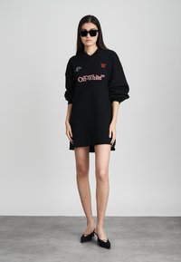 Črna oversize kapučinasta obleka s polo ovratnikom, ki na sprednji strani nosi rdeči logo "Off-White™". Model nosi kvadratna sončna očala in črne petke.