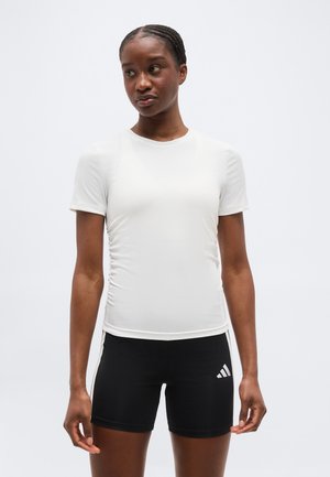 Γυναίκα με πλεξούδες που φοράει εφαρμοστό λευκό κοντομάνικο μπλουζάκι και μαύρο σορτς Adidas, στέκεται μπροστά σε απλό φόντο.