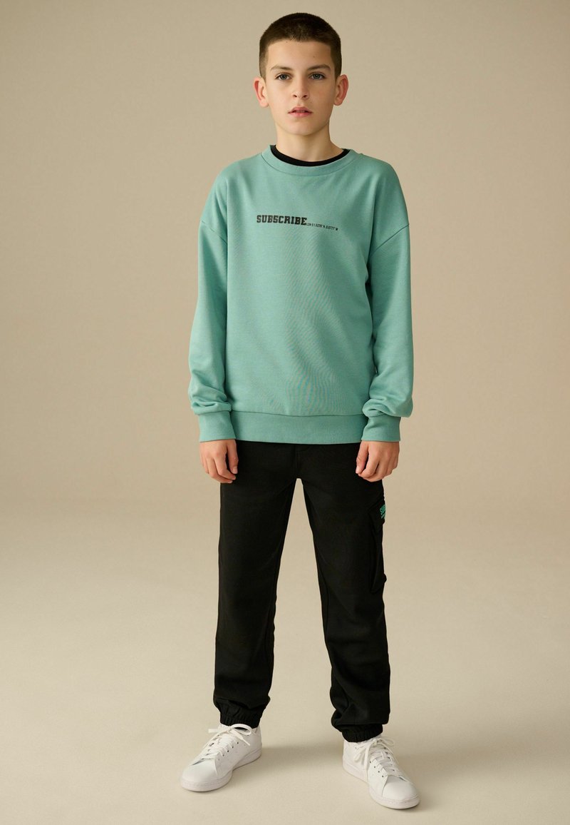 Mintgrøn sweatshirt med sort tekst, der står "ABONNER", parret med sorte joggers og hvide sneakers. Glat tekstur, afslappet pasform.