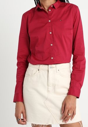 Kobieta ubrana w czerwoną koszulę na guziki z długim rękawem, wpuszczoną w wysokowatą, kremową spódnicę jeansową z postrzępionym dołem.