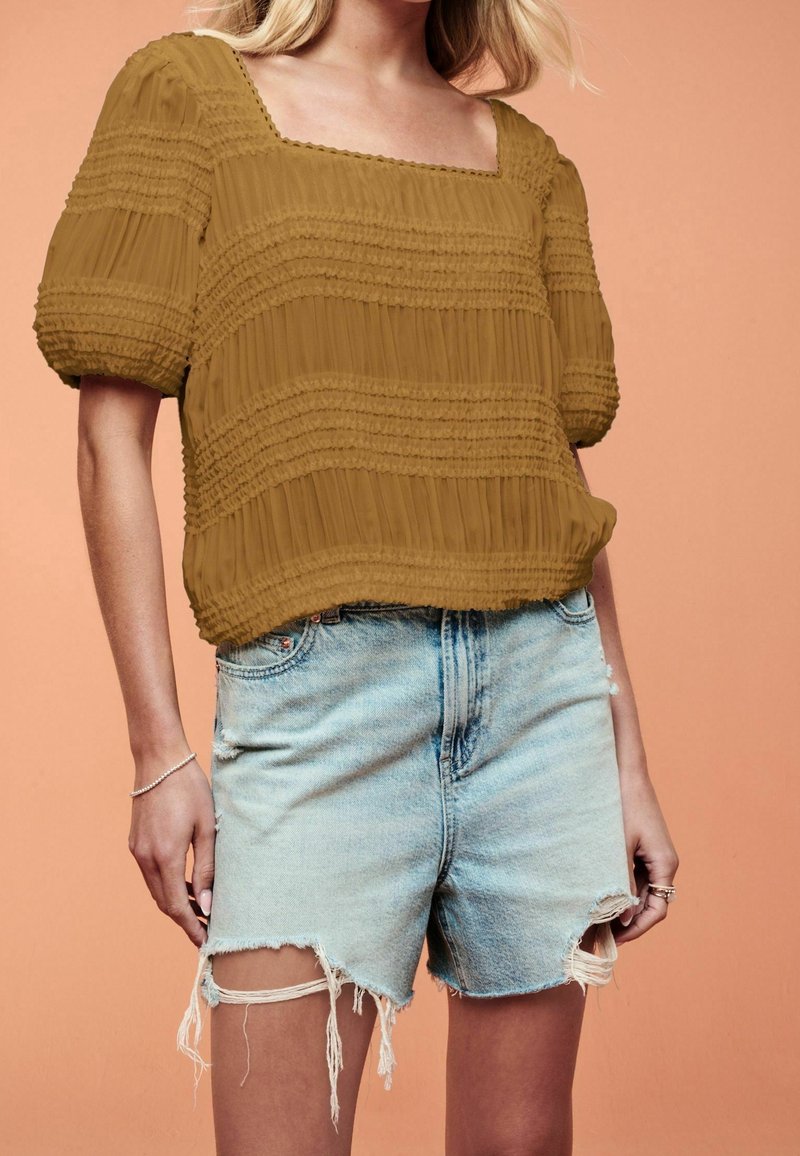 Brun tekstureret bluse med firkantet halsudskæring og puffede ærmer, parret med lyse denim shorts med slidte detaljer.