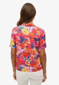 Chemise à manches courtes florale rouge, ornée de diverses fleurs colorées et de feuilles, avec une texture douce et une coupe décontractée. Col arrière visible.