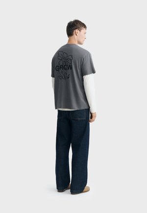 Homme portant une chemise blanche à manches longues superposée avec une chemise grise à manches courtes avec "GROW" et un motif floral dans le dos, un jean large foncé et des chaussures marron.