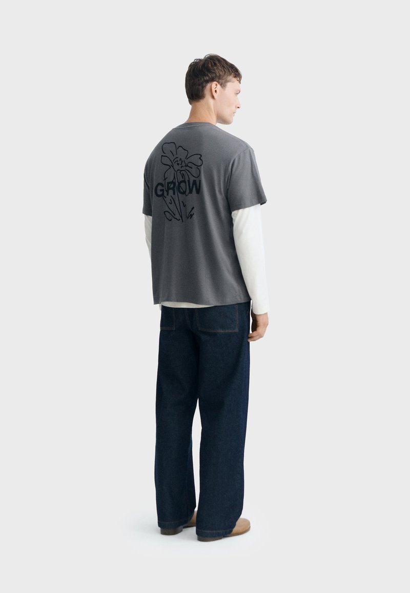 Homme portant une chemise blanche à manches longues superposée avec une chemise grise à manches courtes avec "GROW" et un motif floral dans le dos, un jean large foncé et des chaussures marron.