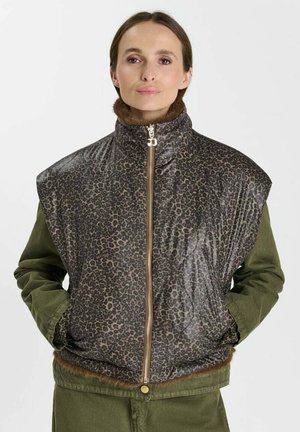Frau trägt eine gesteppte Weste mit Leopardenmuster und grünen langen Ärmeln, Hände in den Taschen, steht vor einem schlichten weißen Hintergrund.