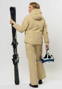 Eine Person in einer braunen Skijacke und -hose hält in einer Hand ein Paar Skier vertikal und in der anderen einen weißen Skihelm mit Skibrille.