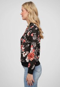 Veste bomber noire à fleurs, ornée de fleurs rouges et blanches, avec des poignets et un ourlet côtelés, associée à un jean bleu clair.