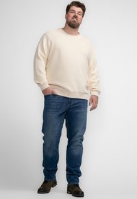 Man staat met één hand in zijn zak, draagt een crèmekleurige sweatshirt, blauwe jeans en bruine laarzen tegen een effen grijze achtergrond.