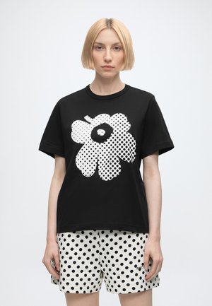 Kvinde med kort blondt hår iført en sort t-shirt med et stort hvidt blomsterprint og hvide shorts med sorte prikker.