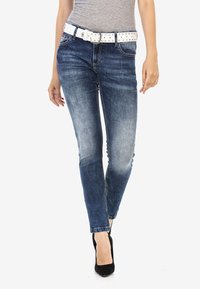 Jean skinny bleus avec une texture délavée et une ceinture blanche cloutée, assortis à des chaussures à talons hauts noires, jambes du mannequin sur un fond neutre.