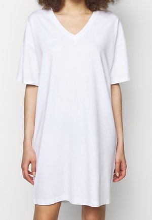 Personne portant une robe blanche ample à manches courtes et col en V, debout devant un fond clair uni.