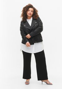 Blouson en cuir noir sur une chemise blanche, associé à un pantalon noir à jambes larges et des sandales à motifs avec des lanières. Cheveux bouclés.