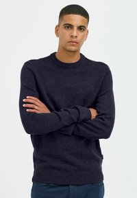 Pull marin noir à col rond, à manches longues, avec une texture tricotée, des poignets et un ourlet côtelés, associé à un pantalon bleu.