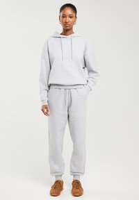 Kobieta stojąca, ubrana w jasnoszarą bluzę z kapturem i dopasowane spodnie dresowe oraz brązowe trampki, na jednolitym tle.