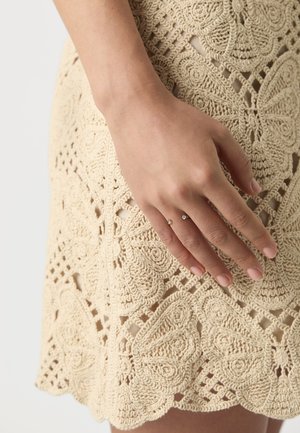 Abito all'uncinetto beige con intricati motivi a farfalla e orlo a onde, abbinato a una mano che indossa un delicato anello con due piccole pietre.