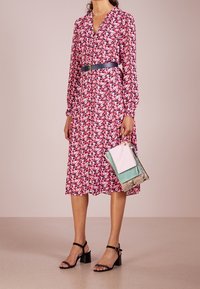 Abito midi floreale in tessuto rosso e bianco, con chiusura frontale a bottoni e maniche lunghe, segnato in vita da una cintura nera. Indossato con tacchi neri.
