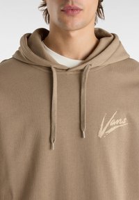 Sweat à capuche kaki en tissu doux avec une poche avant, des cordons de serrage et un logo crème sur le côté gauche de la poitrine.