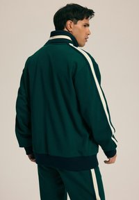 Zielona sportowa kurtka z białymi paskami, z wysokim kołnierzem i ściągaczami przy mankietach. Wykonana z miękkiego, teksturowanego materiału.
