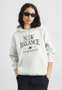 Ανοιχτό γκρι φούτερ με κουκούλα με εκτυπωμένα τα λόγια "NEW BALANCE" και "ATHLETICS" σε μαύρο, διαθέτει μπροστινή τσέπη και κορδόνια. Το μοντέλο φοράει μαύρα γυαλιά ηλίου.