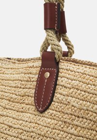 Sac fourre-tout en fibres naturelles tressées avec une texture paille beige, des lanières en cuir marron fixées par des rivets en laiton, et des poignées en corde torsadée.